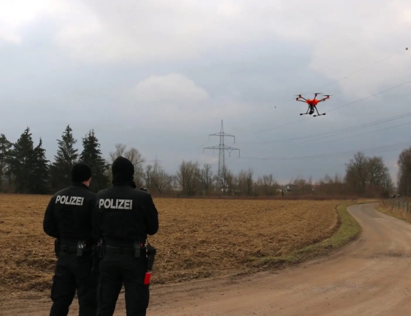 Zwei Polizist*innen stehen vor einem gepflügten Feld, vor ihnen fliegt ein Multicopter/Drohne.